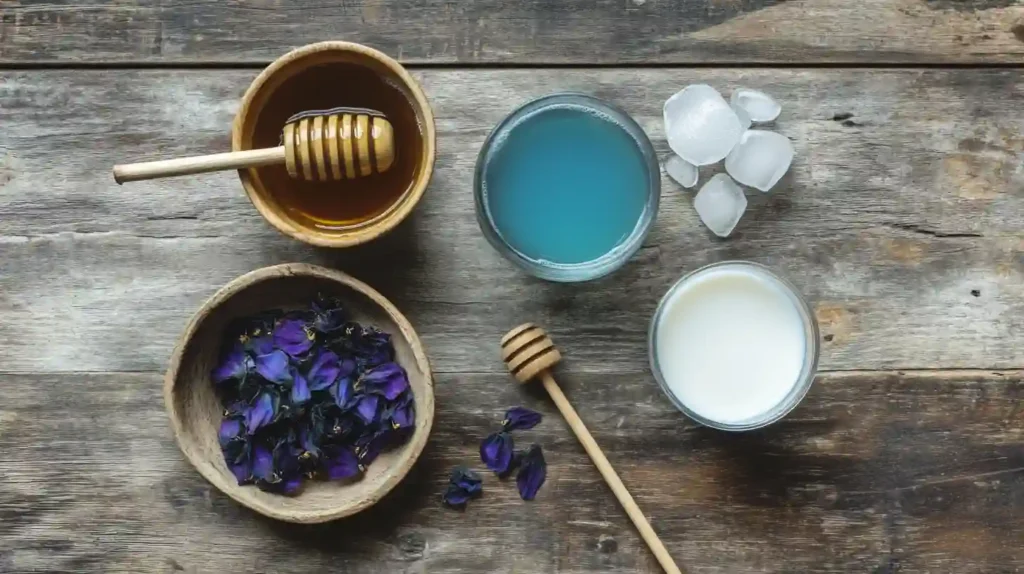A top-down view of ingredients for Butterfly Pea Milk Tea, including dried butterfly pea flowers, blue tea, milk, honey, ice, and a bamboo straw on a wooden surface.
